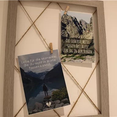 Two postcards hang on a frame. The left card shows a person by the shore of a lake, while the right card presents an inspiring quote in nature.