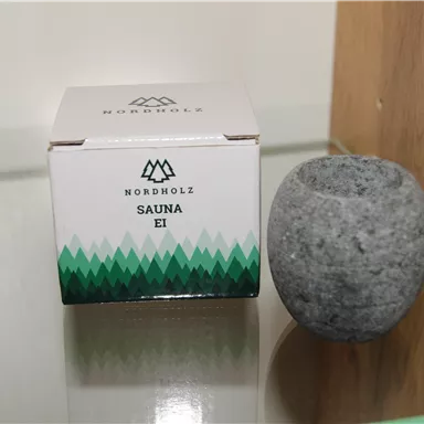 A packaging with the label "Sauna Egg" is next to a round, gray stone object. Both items are placed on a smooth surface.
