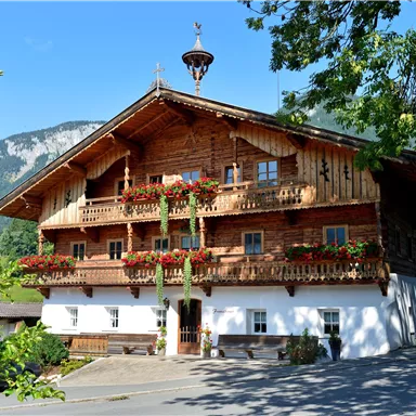 A traditional wooden house with flower-adorned balconies. In the background, green mountains and blue sky can be seen.