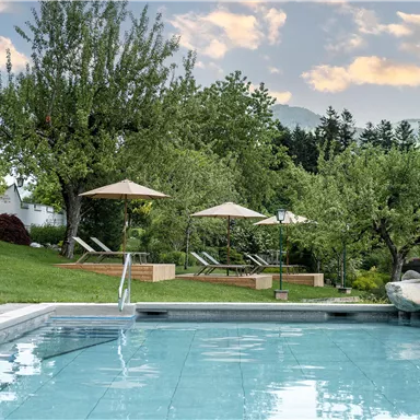 Ein schöner Pool umgeben von gepflegtem Grün und Bäumen. Entspannte Liegen und Sonnenschirme laden zum Verweilen ein.