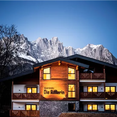 Ein charmantes Holzhaus mit warm beleuchteten Fenstern. Im Hintergrund erheben sich majestätische Berge unter einem klaren Himmel.
