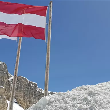 Eine österreichische Fahne weht vor einem klaren blauen Himmel. Im Vordergrund ist ein großer Haufen Schnee zu sehen.