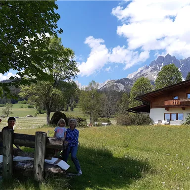 A group of people stands in a meadow in front of a traditional house in the mountains. In the background, green hills and impressive peaks can be seen under a blue sky with a few clouds.