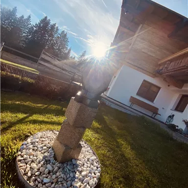 Ein schöner Garten mit einem hölzernen Haus im Hintergrund. In der Mitte steht eine dekorative Skulptur auf Kieselsteinen, während die Sonne im Hintergrund scheint.