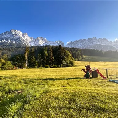 A vast meadow with a playground and impressive mountains in the background. The sky is clear and blue, the landscape appears idyllic.