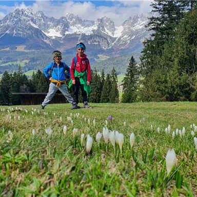 Zwei Kinder stehen auf einer blühenden Wiese mit Schneeglöckchen. Im Hintergrund sind majestätische Berge und eine klare blaue Himmel sichtbar.