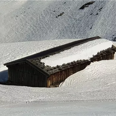 Eine alte Holzhütte, die teilweise von Schnee bedeckt ist. Sie steht in einer verschneiten Landschaft mit sanften Hügeln.