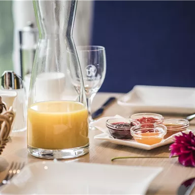 Ein Tischgedeck mit einem Krug Orangensaft, einem Korb mit Brötchen und verschiedenen Marmeladen. Neben dem Tisch stehen Gläser und eine Blume.