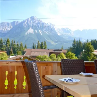 A beautiful terrace with a wooden table and chairs. In the background, impressive mountains and a blue sky can be seen.