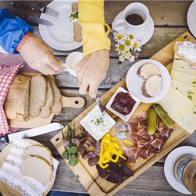 A laid table with various foods, including bread, cheese, sausage, and pickled vegetables. There are also coffee cups and a colorful salad.