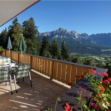 Eine sonnige Terrasse mit Blick auf die Berge. Tische und Stühle laden zum Entspannen ein, umgeben von grünen Bäumen und bunten Blumen.