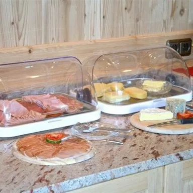 A buffet with various types of sausages and cheeses presented under plastic covers. Next to the buffet, there are plates and cutlery on a wooden panel.