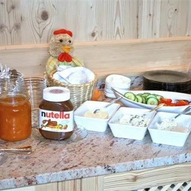 A breakfast table with various jams, Nutella, and prepared plates. Fresh vegetables like cucumbers and bell peppers are also visible next to a decorative figure.