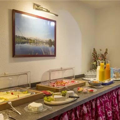 A breakfast buffet with various types of cheese, cold cuts, and fruit. In the background, orange juice and a pretty picture on the wall can be seen.