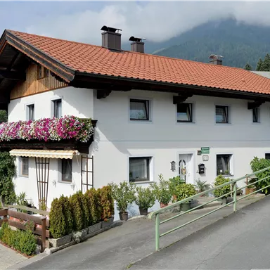 Ein charmantes Haus mit einem roten Ziegeldach und bunten Blumen auf dem Balkon. Im Hintergrund sind sanfte Hügel und eine neblige Berglandschaft zu sehen.