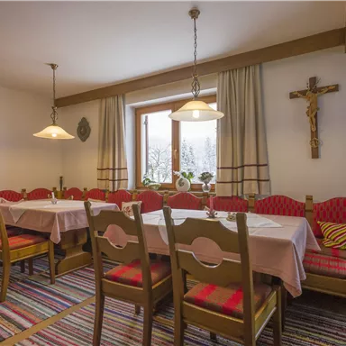 Ein gemütlicher Speisesaal mit langen Tischen und roten gepolsterten Stühlen. An der Wand hängt ein Kreuz und große Fenster lassen viel Licht herein.