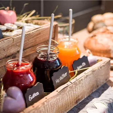 Three glass jars with different jams are placed in a wooden box. In the background, fresh bread and other treats can be seen.