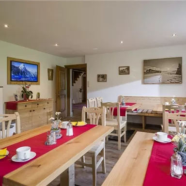 A cozy dining room with wooden tables and red tablecloths. In the background, there is a fireplace and decorative pictures on the wall.