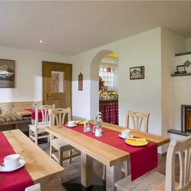 An inviting dining area with wooden chairs and tables. The tables are set with coffee cups and red napkins, while a fireplace adds coziness.