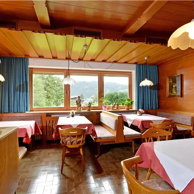 Eine gemütliche Stube mit Holzmöbeln und bunten Tischdecken. Große Fenster bieten einen Blick ins Grüne und lassen viel Tageslicht herein.