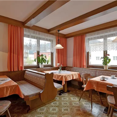 Ein gemütlicher Gastraum mit Holzmöbeln und orangen Tischdecken. Die Fenster sind mit Vorhängen dekoriert, und es stehen Pflanzen auf den Tischen.