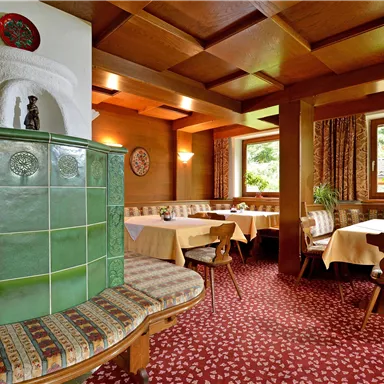 Ein gemütliches Restaurant mit hellem Holzinterieur und traditionellen Tischen. Ein schöner Kachelofen und Fenster, die viel Tageslicht hereinlassen.