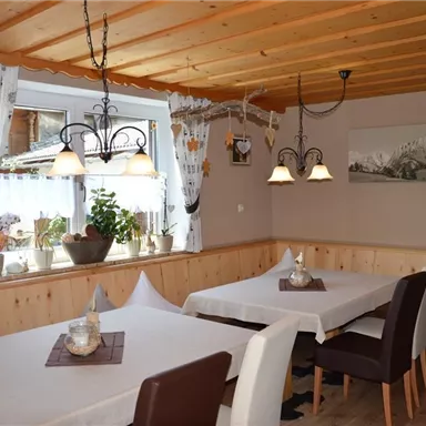 A cozy dining area with wooden paneling and large windows. The tables are simply set, and the walls are decorated with pictures and plants.