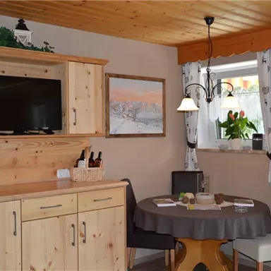 A cozy kitchen with wooden cabinets and a table. A TV hangs on the wall and a window with curtains lets in natural light.