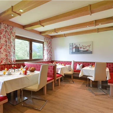 A cozy dining room with red benches and tablecloths. The windows offer a view of the greenery and the room is warmly decorated.