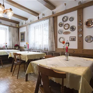A cozy restaurant with wooden furniture and decorative plates on the walls. The tables are covered with white tablecloths and each is adorned with a flower.