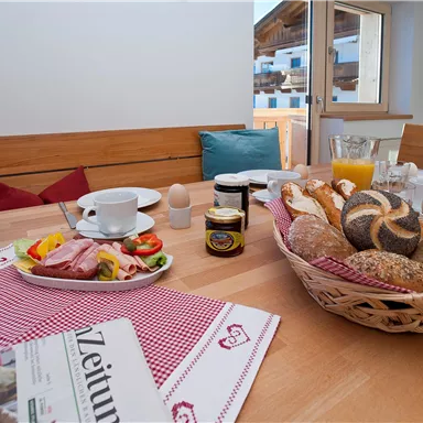 Ein gemütlicher Frühstückstisch mit frischen Brötchen, Wurst, Käse und Obst. Ein Blumenstrauß und eine Zeitung runden das Bild ab.