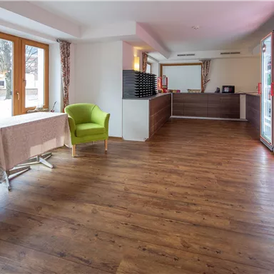 A modernly designed reception area with a bright wooden floor. Two green chairs are in front of large windows, and the reception desk is visible in the background.