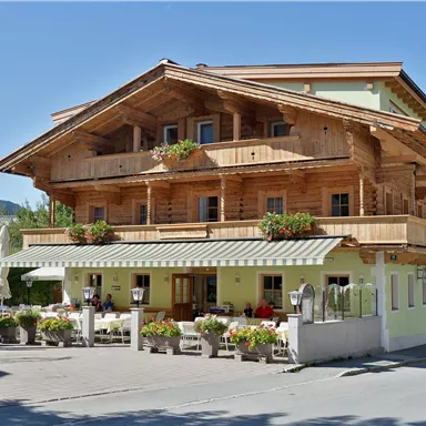 Ein hübsches Holzhaus im alpinen Stil mit Balkon und Blumentöpfen. Vor dem Gebäude sind Tische und Stühle sowie farbige Sonnenschirme aufgestellt.