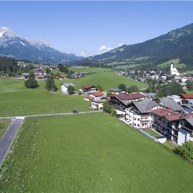 A picturesque landscape with green meadows and small houses. In the background, mountains and a clear sky can be seen.