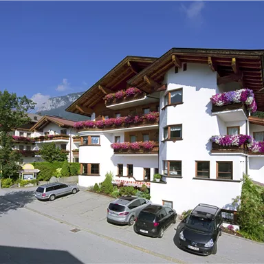 A well-maintained building in Alpine style with colorful flower balconies. Surrounded by trees and a quiet street with parked cars.