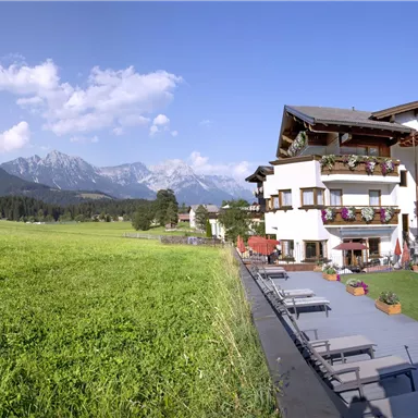 A picturesque hotel with balconies, surrounded by green meadows and mountains in the background. The sky is clear and blue, perfect for a relaxing stay.