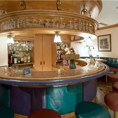 A cozy bar with a round counter and various seating options. In the background, there are shelves with drinks and subdued lighting.
