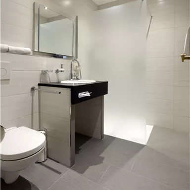 A modern bathroom with a black sink, a mirror, and a shower cabin. The walls are light tiled and the floor is gray.