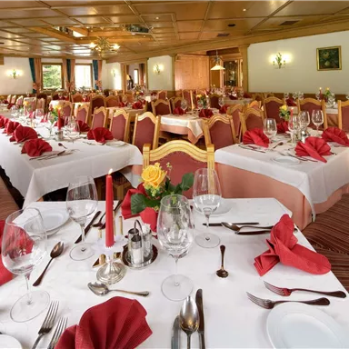 An elegant dining room with many set tables. Red napkins and floral arrangements create a festive atmosphere.