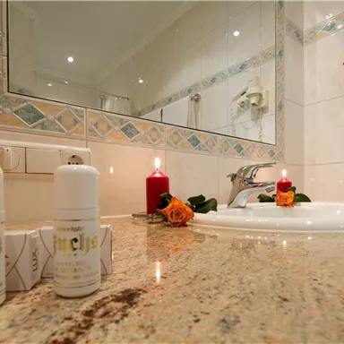 A bathroom with a white sink and a large mirror. On the shelf are two bottles as well as a candle and a rose decorated.