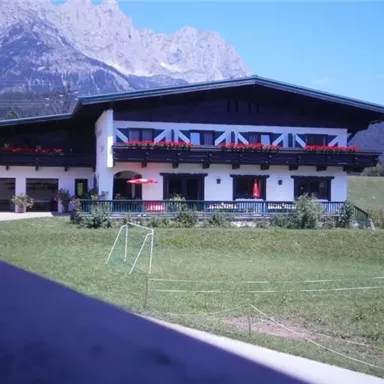 A traditional Alpine building with balconies and red flower boxes. In the background, mountains and a green field can be seen.