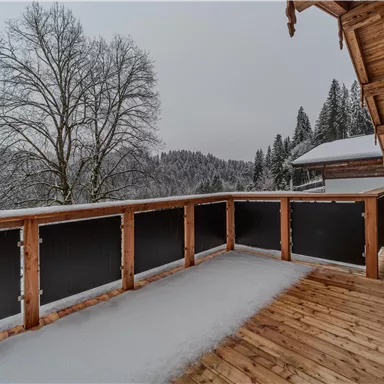A balcony with a wooden railing and a view of a snowy landscape. In the background, trees and hills are visible.