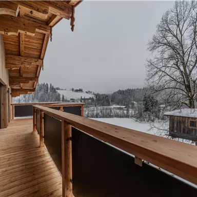 A wooden balcony overlooking a snowy landscape. In the background, trees and a wooden house are visible.