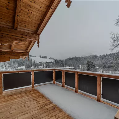 A cozy balcony with a wooden railing and a view of snow-covered mountains. The surroundings are quiet and wintry.