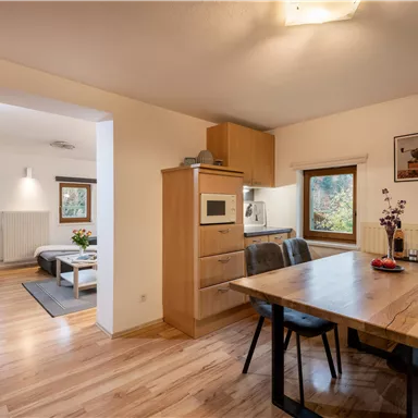 Eine moderne Küche mit einem großen Holztisch und stilvollen Stühlen. Im Hintergrund sind gemütliche Wohnbereiche mit Fenstern, die Tageslicht hereinlassen.