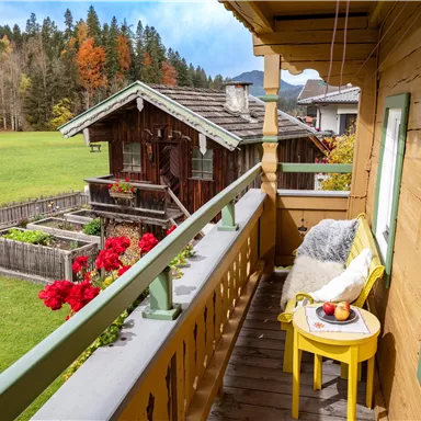 Ein Balkon mit einer Sitzgelegenheit, die eine Aussicht auf den Garten und das alte "Waschhaus" bietet. Bunte Blumen und Bäume im Hintergrund sorgen für eine gemütliche Atmosphäre.