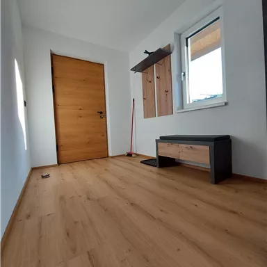 A bright hallway with a wooden floor and a large door.
On the wall, there are coat hooks and a small bench.
