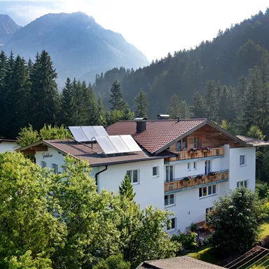 A modern house in the mountains, surrounded by green trees and mountains. The solar panels on the roof showcase an eco-friendly design.