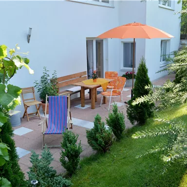 A cozy terrace with a wooden table and chairs under an orange parasol. Surrounded by green plants and lawn, the spot is ideal for relaxation.