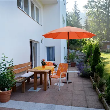 A cozy outdoor area with a table, chairs, and an orange parasol. Surrounded by plants and a well-maintained garden.